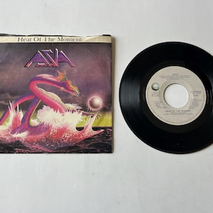 May include: A 7-inch vinyl record by the band Asia. The record is black with a white label that says "Asia" and "Heat Of The Moment". The cover art features a purple and pink dragon-like creature emerging from a wave.