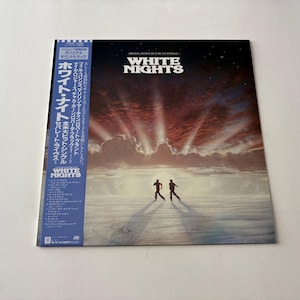 Puede incluir: Portada de álbum de vinilo de la película "White Nights". La portada presenta un cielo dramático con brillantes rayos de luz y dos figuras silueteadas. El título del álbum está en letras blancas grandes. Una pegatina azul con texto japonés está a la izquierda.