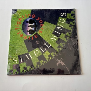 Peut inclure: Vinyle Simple Minds, intitulé "Street Fighting Years", encore sous blister. La pochette présente un motif géométrique vert et noir, avec le nom du groupe et le titre de l'album en blanc et rouge. Une image circulaire montre des silhouettes.