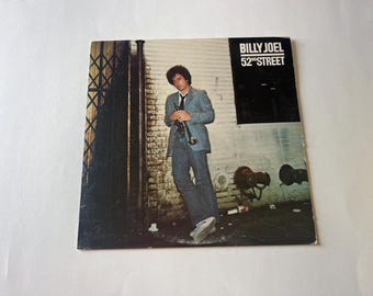 May include: A vintage vinyl record album cover featuring Billy Joel. The cover art shows a man in a blue suit and jeans standing in an alleyway. The album title "52nd Street" is printed in white on a black background.