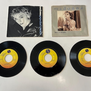 May include: Three black vinyl 45 rpm records with yellow labels featuring Madonna's "Like a Virgin", "Papa Don't Preach", and "Vogue" singles. The records are displayed on a white surface.  The image also includes two Madonna album covers, "Like a Virgin" and "Papa Don't Preach".