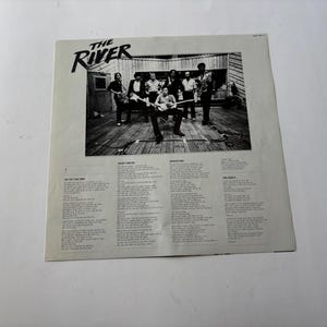 May include: Black and white album cover for "The River" featuring a band performing on a wooden stage. The album title is in a stylized font. Lyrics are printed below the image.