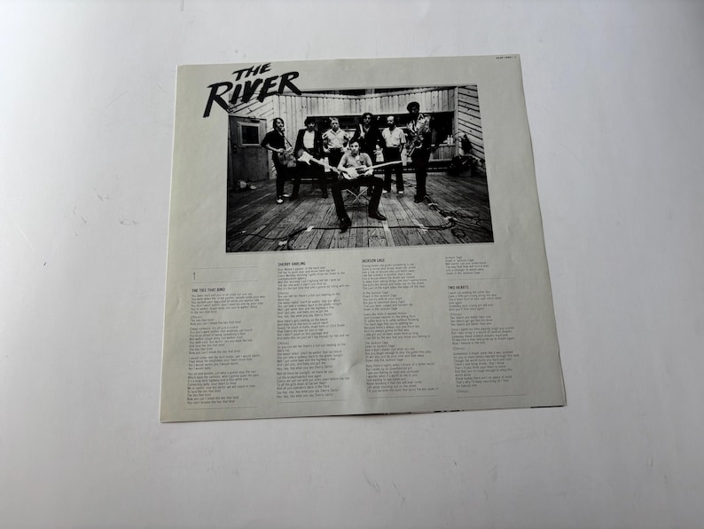 May include: Black and white album cover for "The River" featuring a band performing on a wooden stage. The album title is in a stylized font. Lyrics are printed below the image.