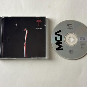 May include: A compact disc of Steely Dan's album "Aja" with the MCA logo. The album cover is black with a vertical red and white design. The CD is silver with black text.