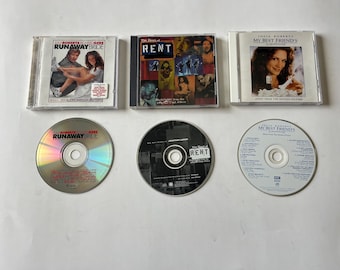 May include: Three CD cases and three CDs, all featuring movie soundtracks. The cases are white with colorful images and text. The CDs are silver with printed labels. The titles are "Runaway Bride", "Rent", and "My Best Friend's Wedding".