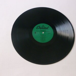 Op de afbeelding: Zwarte vinylplaat met een groen label waarop "Capitol" en "The Miracles" staat met een lijst van nummers. De plaat ligt op een wit oppervlak.