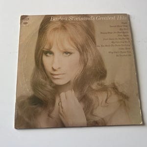 May include: A vintage vinyl record cover featuring a black and white photo of Barbra Streisand. The cover is titled "Barbra Streisand's Greatest Hits" and lists the songs included on the record: "People", "Second Hand Rose", "My Son", "My Man", "Happy Days Are Here Again", "Free Again", "Don't Rain On My Parade", "My Coloring Book", "You Make Me Feel So Young", "Gotta Move", "Why Did I Choose You", "He Touched Me".