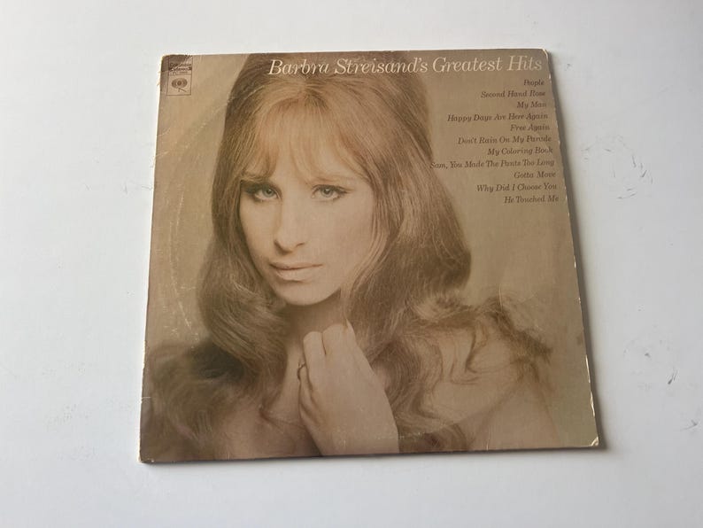 May include: A vintage vinyl record cover featuring a black and white photo of Barbra Streisand. The cover is titled "Barbra Streisand's Greatest Hits" and lists the songs included on the record: "People", "Second Hand Rose", "My Son", "My Man", "Happy Days Are Here Again", "Free Again", "Don't Rain On My Parade", "My Coloring Book", "You Make Me Feel So Young", "Gotta Move", "Why Did I Choose You", "He Touched Me".