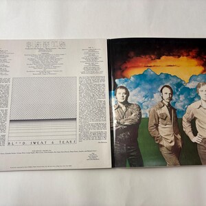 May include: An open vinyl record album featuring the band Blood, Sweat & Tears. The album cover shows three men in front of a colorful sky. The album's inner sleeve has text and a dotted pattern.
