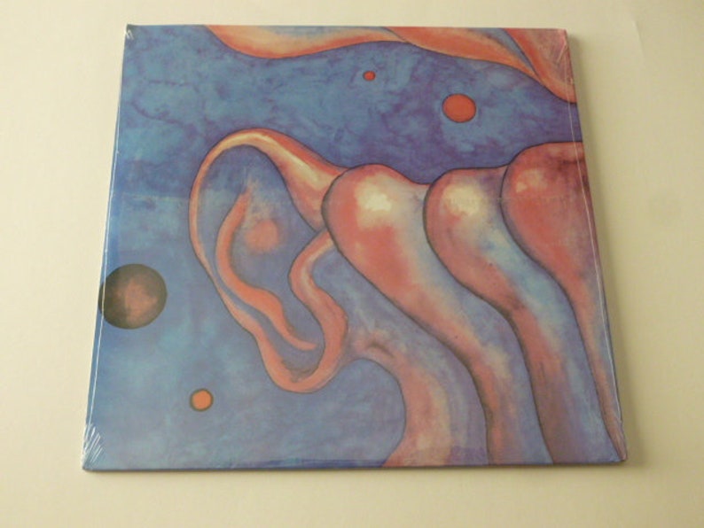 May include: A vinyl record cover with a blue and pink abstract design. The design features a large, pink, organic shape in the center of the cover. The shape is surrounded by smaller, blue and pink shapes. The cover also features a few small, red circles.