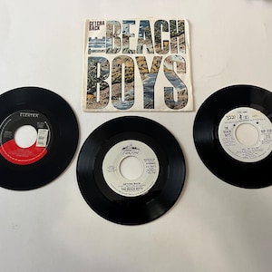 May include: Three black vinyl records with white labels. The Beach Boys album cover is visible in the background. The album cover is white with blue and green text and images. The album title is "The Beach Boys" and the song title is "Getcha Back".
