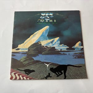Peut inclure: Une pochette de disque vinyle carrée pour l'album Drama du groupe Yes. L'illustration présente un paysage bleu foncé avec de grands icebergs blancs, un plan d'eau et les silhouettes noires de trois chats au premier plan sur une surface texturée.