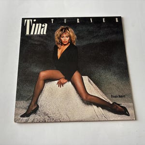 May include: Album cover featuring Tina Turner in a black outfit, fishnet stockings, and heels. The album title "Tina" is at the top left, with "Private Dancer" at the bottom right. The background is a dark, textured gradient.