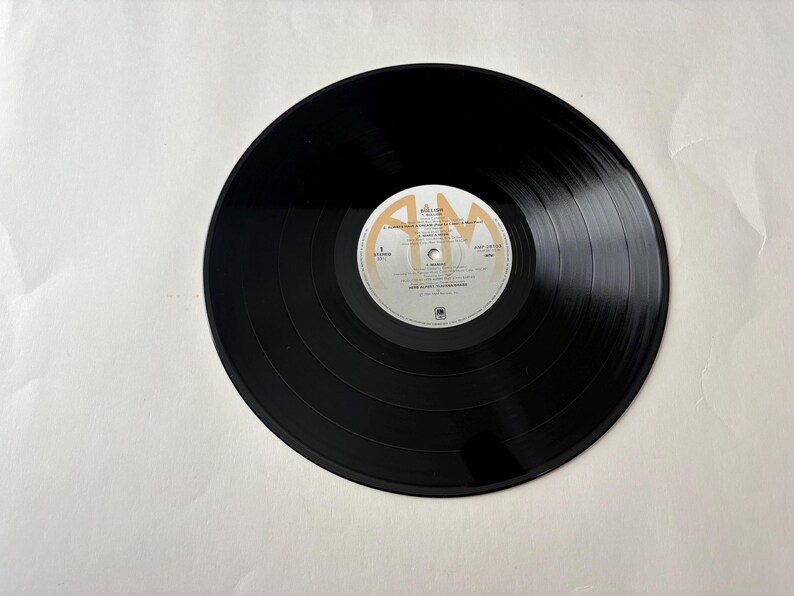Puede incluir: Un disco de vinilo negro con una etiqueta blanca y dorada. El disco tiene surcos conc&eacute;ntricos y est&aacute; centrado en un fondo blanco. La etiqueta presenta texto y un logotipo. Es un art&iacute;culo de m&uacute;sica vintage.