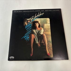 May include: A vinyl record album cover for the movie "Flashdance." The cover features a woman in a gray sweater and red heels. The title "Flashdance" is in a stylized font with blue accents against a black background. The words "Original Soundtrack From The Motion Picture" are at the top.