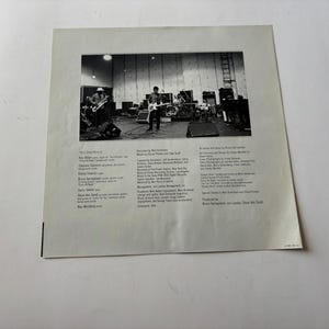 May include: Black and white photograph of a band performing on a stage. The image is a close-up of a record album cover. The album cover has text on the left and right sides of the image.