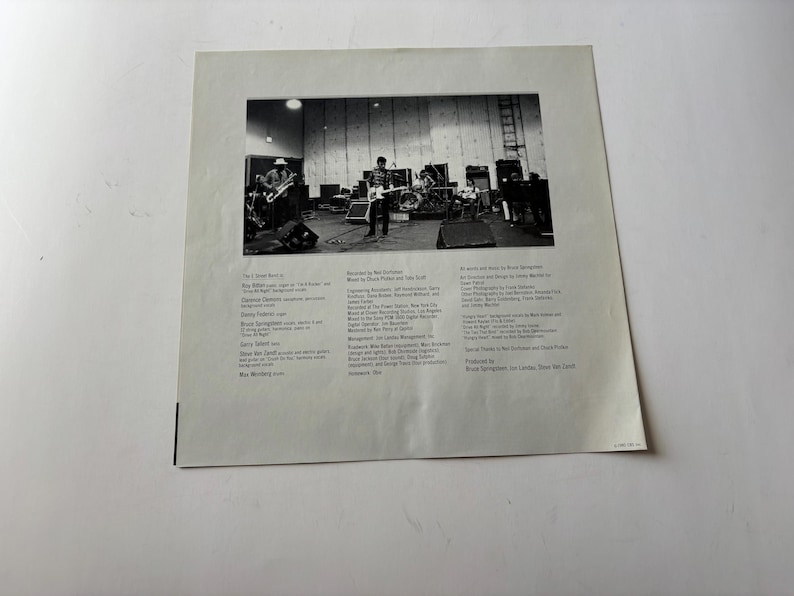 May include: Black and white photograph of a band performing on a stage. The image is a close-up of a record album cover. The album cover has text on the left and right sides of the image.