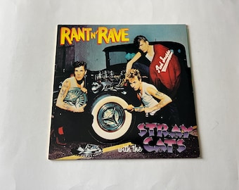 May include: A vintage album cover featuring a band called "Rant 'n Rave with the Stray Cats". The cover features three men in front of a hot rod car. The men are wearing leather jackets and jeans. The car is red and black and has a large chrome grill.