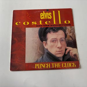 May include: Album cover for "Punch the Clock" by Elvis Costello and The Attractions. The cover features a portrait of a man with glasses and a hat, set against a red background with yellow text.