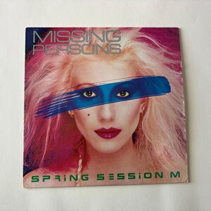 May include: Album cover for Missing Persons' "Spring Session M." The image features a woman with pink hair, red lipstick, and a blue stripe across her eyes. The band's name is in silver letters, and the album title is in green.