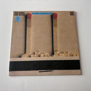 May include: Album cover for "One on One" by Bob James and Earl Klugh. The cover features a brown background with two large matchstick designs. The match heads are red, and the album title is in blue.