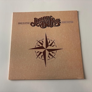 Pode incluir: Uma capa de álbum vintage com o texto "Jimmy Buffett" em uma fonte marrom estilizada. Abaixo do texto, há um desenho de rosa dos ventos marrom. O título do álbum "Changes in Latitudes, Changes in Attitudes" também é visível.