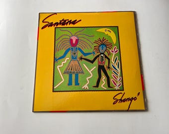 May include: A yellow vinyl record album cover with the text "Santana" in red and "Shango" in black. The cover features a colorful illustration of two figures with a green background.