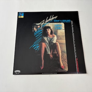 May include: A vinyl record album cover for the movie "Flashdance" featuring Irene Cara. The cover is predominantly black with the title in a stylized font and a photo of Cara. The album includes the song "What a Feeling."