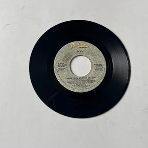 May include: A black vinyl record with a silver label. The label has the text "DEVO" and "THEME FROM DOCTOR DETROIT" printed on it.