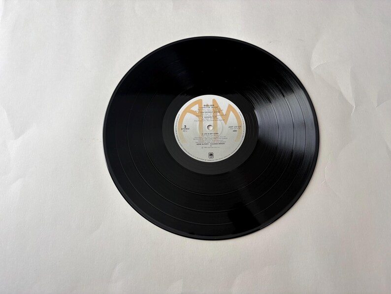 Puede incluir: Un disco de vinilo negro con una etiqueta blanca y dorada. El disco tiene surcos conc&eacute;ntricos y una etiqueta circular con texto. La etiqueta presenta el texto "A&M" y t&iacute;tulos de canciones. El disco est&aacute; sobre un fondo blanco.