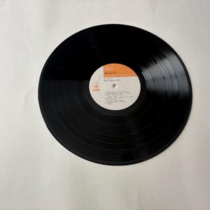 May include: A black vinyl record with a white and orange label. The label features text including the artist's name and song titles. The record has visible grooves and is set against a white background. The record is a vintage music item.