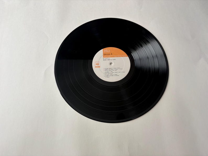 May include: A black vinyl record with a white and orange label. The label features text including the artist's name and song titles. The record has visible grooves and is set against a white background. The record is a vintage music item.