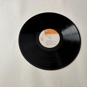 May include: A black vinyl record with a white and orange label. The label includes the artist's name, album title, and a list of songs. The record is centered on a white background.