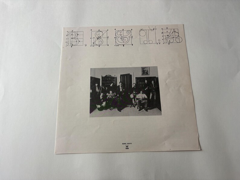 May include: A vintage album cover featuring a black and white photograph of a band in a room. Above the photo are stylized letter designs. The album cover is on a white background.