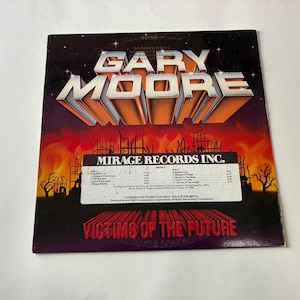 以下が含まれることがあります： ゲイリー・ムーアの「Victims of the Future」のヴィンテージビニールレコードアルバムカバー。アルバムアートは、アーティストの名前を大きな3Dオレンジ色の文字で、燃えるような終末論的なシーンの上に表示しています。 Mirage Records Inc.のラベルが見えます。