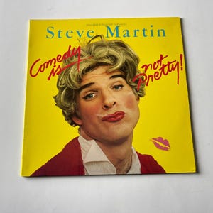 May include: A yellow album cover with the text "Steve Martin Comedy is not Pretty!" in red. A man with blonde hair is wearing a red jacket and a white shirt. He is looking at the camera with a serious expression.