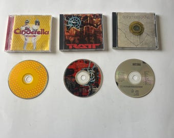 May include: A collection of five music CDs, including albums by Cinderella, Ratt, and Whitesnake. The CDs are arranged in a row on a light brown surface. The CDs are all in jewel cases.