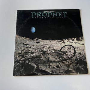 May include: A black vinyl record album cover with the title "PROPHET CYCLE OF THE MOON" in white text. The cover features a dark, moon-like landscape with a small blue planet in the background.