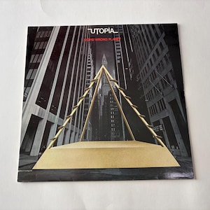 May include: Album cover for "Utopia" by "Oops! Wrong Planet." The artwork shows a gold geometric structure against a black and white cityscape. The album title and artist name are in white and red.