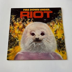 Może przedstawiać: Okładka płyty winylowej do albumu "Fire Down Under" zespołu Riot. Okładka przedstawia białe, futrzane stworzenie z czerwonymi oczami i różową koszulą na żółtym i pomarańczowym tle.
