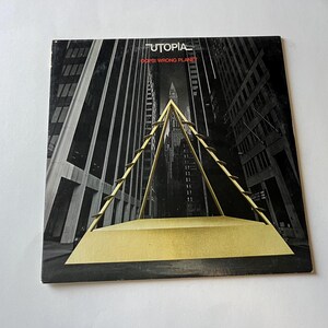 May include: A black and white album cover with the title "Utopia" and the text "Don't  Wrong Planet" in red. The cover features a gold pyramid-shaped structure in front of a city skyline.