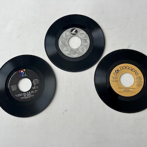 Puede incluir: Tres discos de vinilo negros con diferentes etiquetas. Las etiquetas son doradas y blancas con texto negro. Los discos están sobre una superficie blanca.