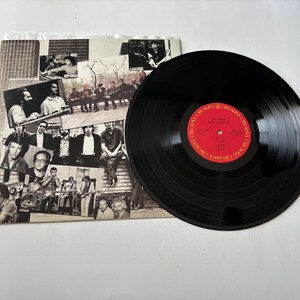 May include: A black vinyl record with a red label featuring white text, next to a record sleeve with black and white images of people. The record is a music album.