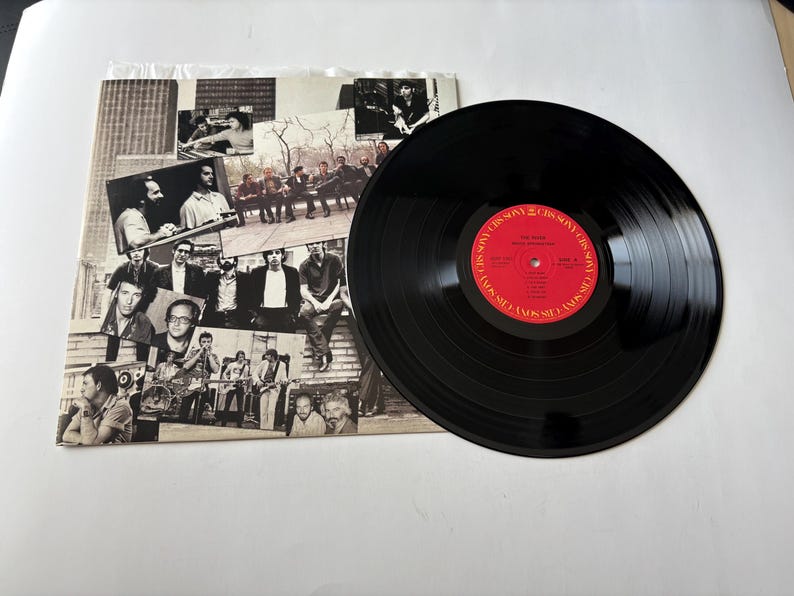 May include: A black vinyl record with a red label featuring white text, next to a record sleeve with black and white images of people. The record is a music album.