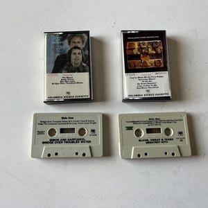 May include: Four cassette tapes, two are white with black text and two are black with white text. The white tapes are labeled "Simon and Garfunkel Bridge Over Troubled Water" and "Blood, Sweat & Tears Greatest Hits". The black tapes are labeled "Bridge Over Troubled Water" and "Blood, Sweat & Tears".