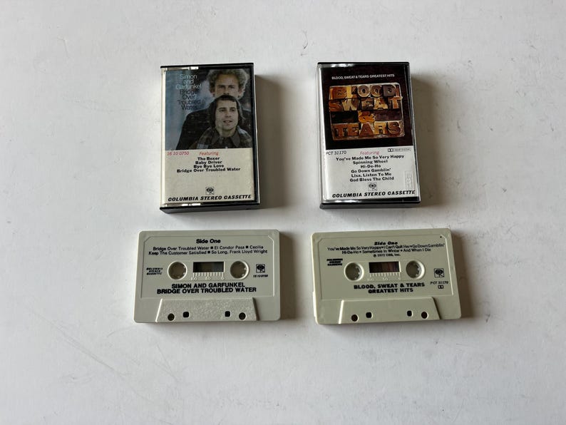 May include: Four cassette tapes, two are white with black text and two are black with white text. The white tapes are labeled "Simon and Garfunkel Bridge Over Troubled Water" and "Blood, Sweat & Tears Greatest Hits". The black tapes are labeled "Bridge Over Troubled Water" and "Blood, Sweat & Tears".