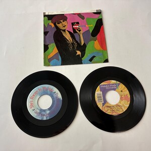 May include: A 7-inch vinyl record set featuring the Raspberry Beret single by Prince and the Revolution. The set includes a colorful illustrated paper sleeve and two black vinyl records with printed center labels displaying track titles.