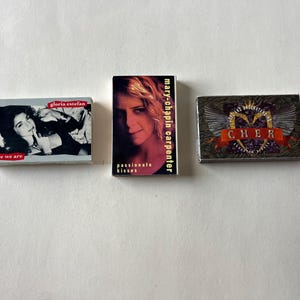 May include: Three cassette tapes featuring different artists. The first tape is by Gloria Estefan and says "Here We Are" on the front. The second tape is by Mary-Chapin Carpenter and says "Passionate Kisses" on the front. The third tape is by Cher and says "Cher" on the front.