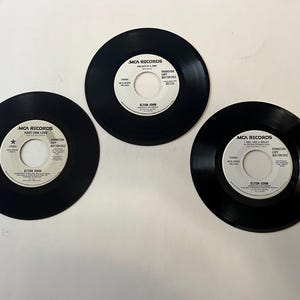 Può includere: Tre dischi in vinile neri con etichette bianche. Le etichette riportano il testo "MCA Records" e il nome dell'artista "Elton John". I titoli delle canzoni sono "One Day At A Time", "Part-Time Love" e "I Feel Like A Bullet".