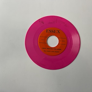 May include: A pink 45 RPM vinyl record with an orange label. The label says "Essex" and "Rock Around The Clock Bill Haley And Comets".